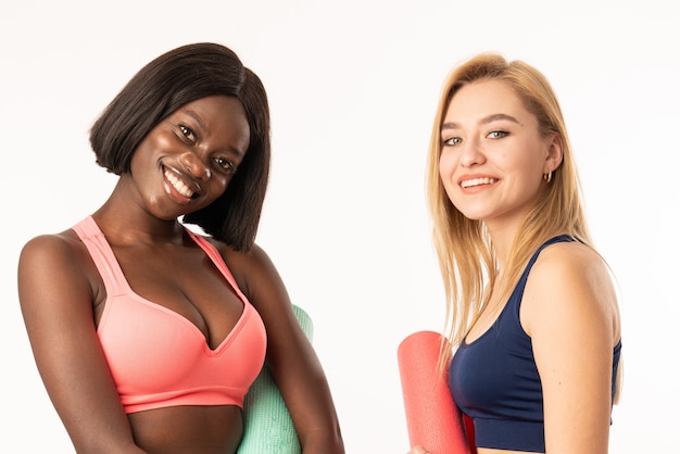 Foto mezzo busto ritratto di due ragazze di fitness affascinante felice sorridente internazionale con materassino yoga