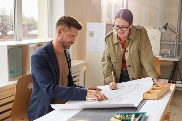 Waist up portrait of two smiling architects pointing at blueprints and discussing work while standing by drawing desk in office lit by sunlight , 