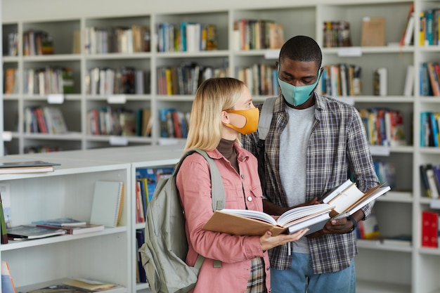 Waist up portrait of two students wearing masks while standing in school library and holding books, 