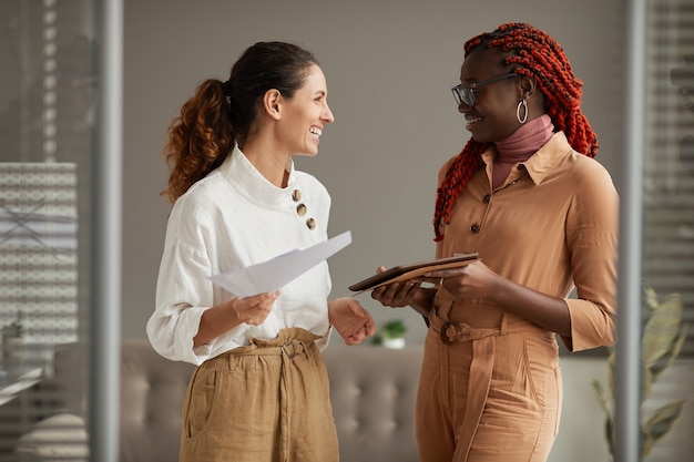 Waist up portrait of two successful young businesswomen smiling cheerfully looking at each other while standing in modern office interior, copy space