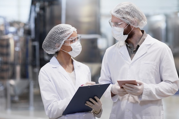 Waist up portrait of two workers wearing protective clothing while discussing production at modern chemical plant