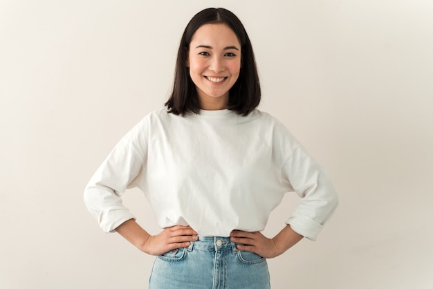 Waist up portrait view of a beautiful happy young woman standing on a white wall and sincerely smiling. Modern girl smiling broadly at camera, while standing against white background