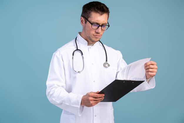 Waist up portrait view of the confident doctor in a uniform and in glasses holding folder and looking at it with serious face. Stock photo