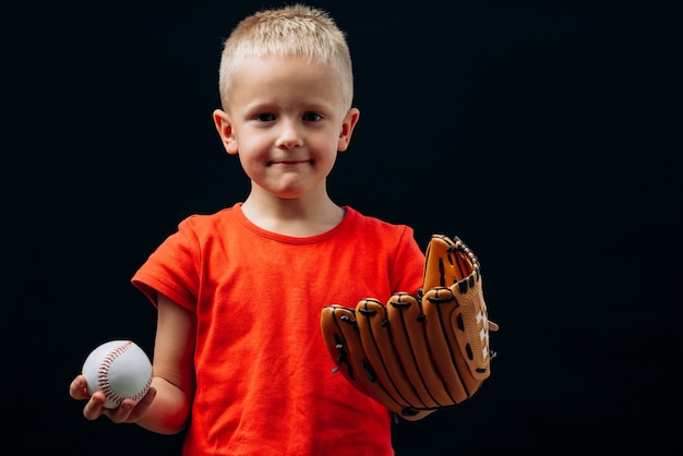 特別な手袋をはめてボールを持ってカメラを見ているかわいい男の子の野球選手のポートレートビューを腰に当てます。黒の背景に分離。子供の頃とスポーツの概念