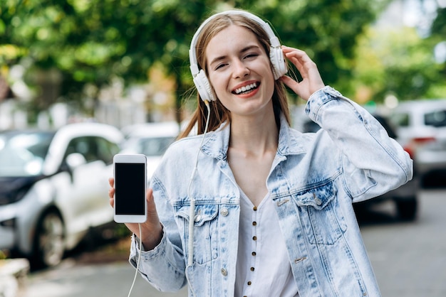 Waist up portrait view of the excited girl laughing and listening music with headphones Woman showing her smartphone in the street