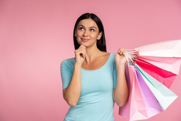 Waist up portrait view of long haired woman holding paper bags and pondering about something. Shopping time, week sale concept. Indoor studio shot isolated on pink background