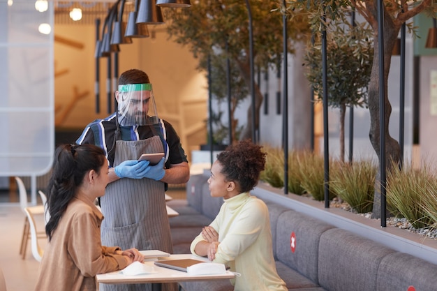 Waist up portrait of waiter wearing face shield and gloves while taking order in cafe with covid safety measures, copy space