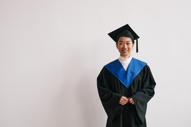 Waist up portrait of young Asian woman wearing graduation robes and smiling at camera, copy space