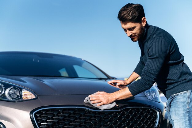 Waist up portrait of a young caucasian man cleaning his car with a microfiber outdoors at sunset while looking at the hood at the street