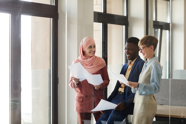 Waist up portrait of young multi-ethnic business team smiling happily and holding documents while enjoying work in office, copy space