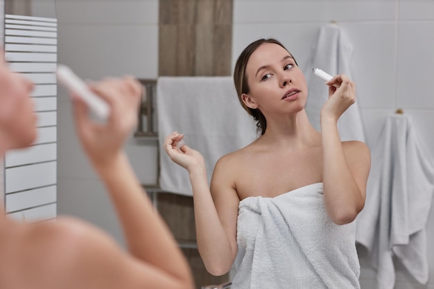 Waist up portrait of young woman using eye cream in bathroom and looking in mirror enjoying self car