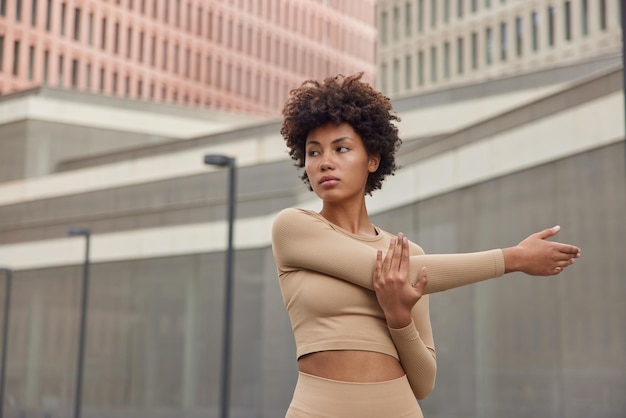 Photo waist up shot of active slim curly haired woman dressed in beige tracksuit stretches arms being in good physical shape concentrated away poses against blurred background prepares for workout