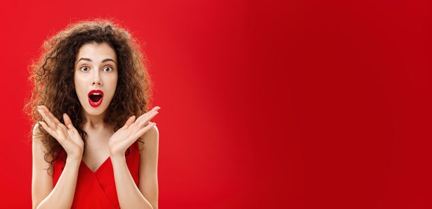 Waist-up shot of amused and astonished good-looking young elegant female with curly hairstyle in red