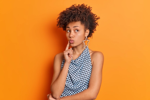 Waist up shot of beautiful curly haired woman keeps lips folded
being deep in thoughts thinks about something wears fashionable t
shirt and star shaped earrings isolated over vivid orange
background