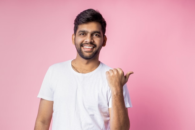 Waist-up shot of cheerful carefree indian man with stubble, wearing white t shirt, pointing left with thumb, laughing, looking at camera, standing over pink background. Advertising concept.