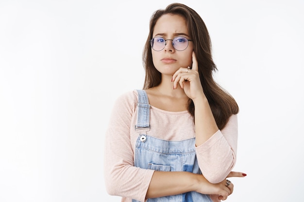 Waist-up shot of concerned smart and beautiful woman in glasses looking at front suspicious and unsure frowning making serious look as hesitating if choice right, expressing disbelief over gray wall.