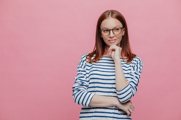 Waist up shot of dreamy thoughtful woman keeps hands partly crossed, holds chin