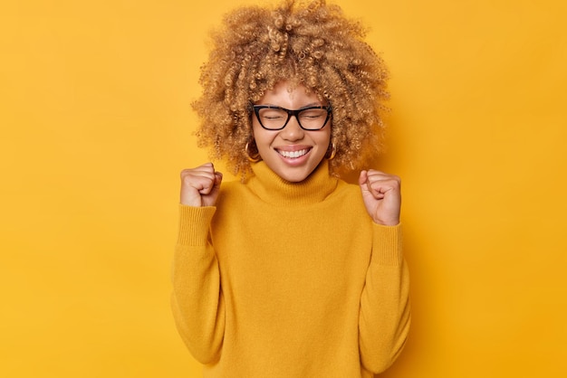 Waist up shot of euphoric positive young woman clenches fists celebrates achievements reaches goal exclaims yes smiles broadly wears eyeglasses casual jumper isolated over yellow background.