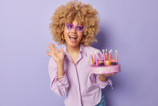 Waist up shot of happy birthday girl with curly blonde hair wears trendy heart shaped sunglasses and shirt holds delicious cake with burning candles isolated over purple background Festive event