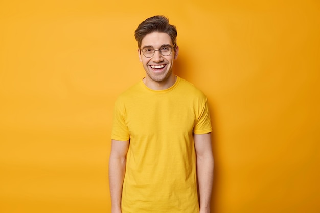 Waist up shot of positive young European man dressed in casual basic t shirt smile happily wears round spectacles poses against vivid yellow background People emotions and feelings concept