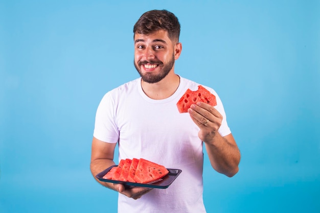 笑顔の男の腰のアップ ショットは、夏の日を楽しんでいます新鮮な熟したスイカのスライスを保持し、おいしい果物を食べたいです青の背景に分離されたカジュアルな白い t シャツを着て、週末にピクニックをしています