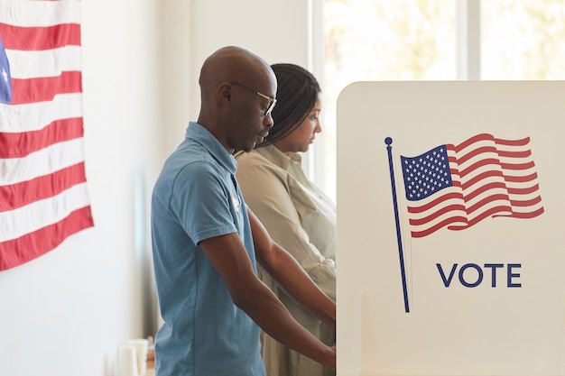 Photo waist up side view portrait of young african-american people standing in voting booth decorated with usa flags, copy space