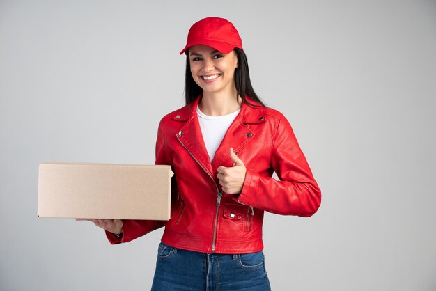 Waist up view portrait of female courier in red uniform handing cardboard parcel to client, carrying parcel with goods ordered online. Indoor studio shot isolated on grey background