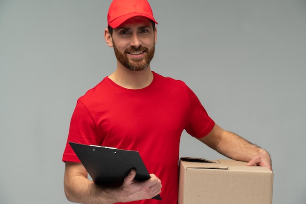 Waist up view portrait of happy male courier in red uniform holding cardboard parcel and looking at the camera with happy emotions Indoor studio shot isolated on grey background