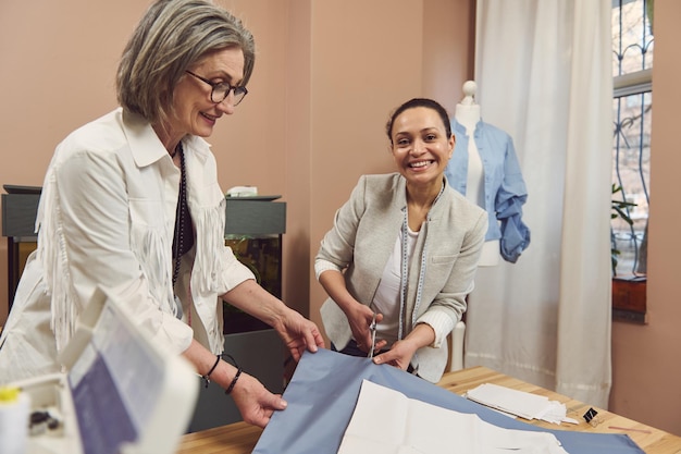 Waistlength portrait of two mature female tailors making clothes patterns on fabric while sewing a classic women's shirt in a fashion design workshop