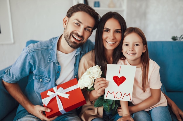 waistup photo of a merry mother sitting between her smiley husband and cute daughter and looking at the camera