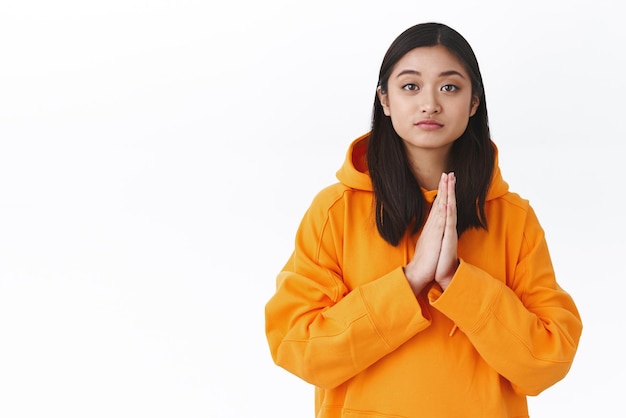 Waistup portrait attractive asian female student in orange hoodie begging team member lend hand asking for help or advice clasp hands together in pray plead gesture white background