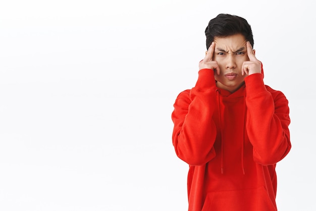 Waistup portrait of funny asian man trying to use telepathy read minds or move objects with power of mind frowning stare intense and squinting holding temples concentrating white background