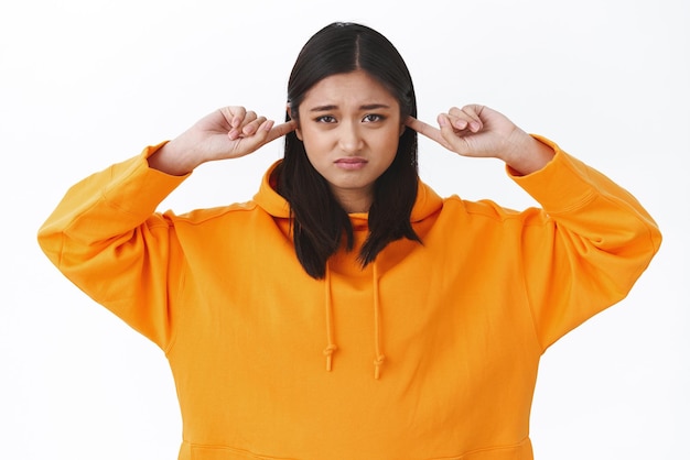 Waistup portret somber en mokkend jonge aziatische vrouw in oranje hoodie grimassen en tuitende lippen gesloten oren niet horen hinderlijk geluid geïrriteerd door lawaai witte achtergrond