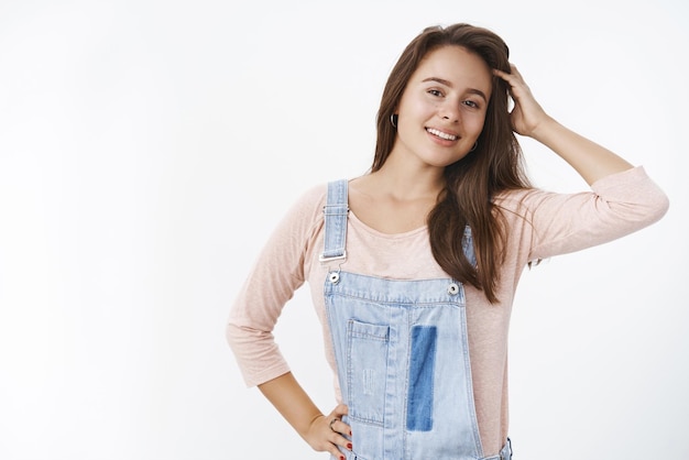 Colpo in vita di una giovane donna bruna sognante e felice che tocca i capelli deliziati sorridendo con i denti bianchi perfetti alla telecamera in piedi soddisfatto del bel taglio di capelli in posa davanti allo specchio sul muro grigio