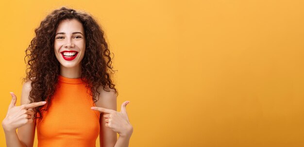 Waistup shot of proud and happy outgoing charming woman with curly hair and red lipstick pointing at