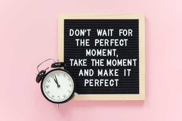 Don't wait for the perfect moment, take the moment and make it perfect. Motivational quote on letter board, black alarm clock