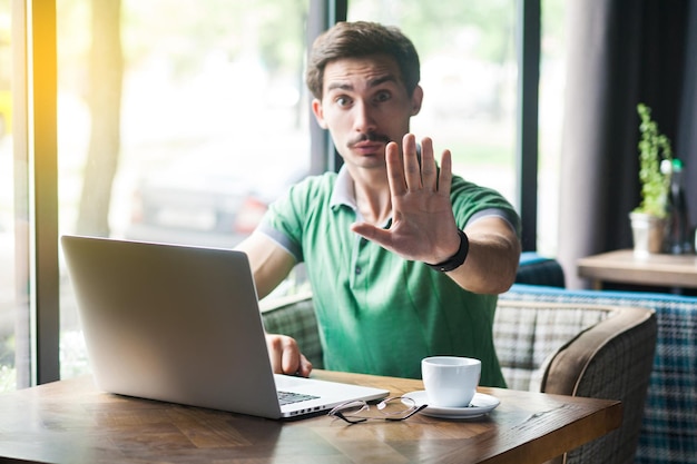 Wait, stop. young serious businessman in green t-shirt sitting,\
working on laptop, looking at camera, showing stop sign gesture.\
business and freelancing concept. indoor shot near window at\
daytime.