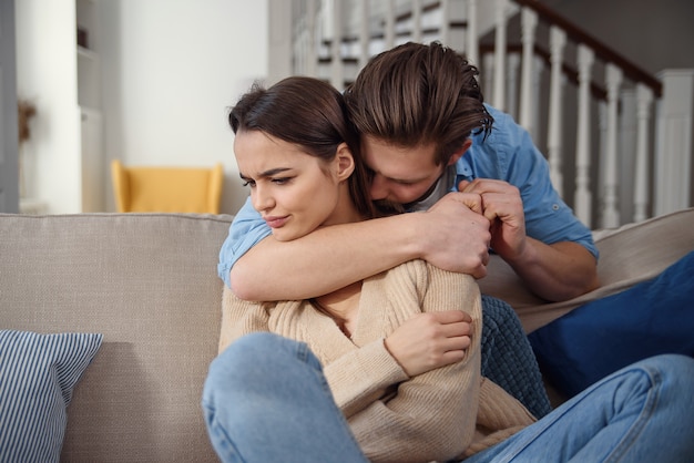 Wait. Worried young man is consoling his girlfriend while touching her arm gently. Woman is holding mobile phone and looking at boyfriend with offence