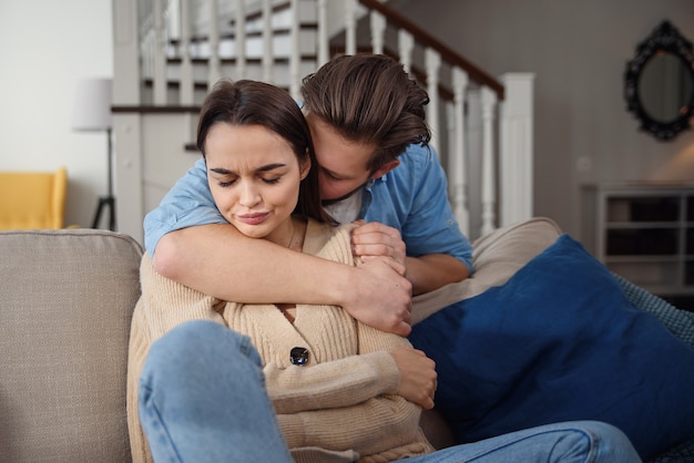 Wait. Worried young man is consoling his girlfriend while touching her arm gently. Woman is holding mobile phone and looking at boyfriend with offence