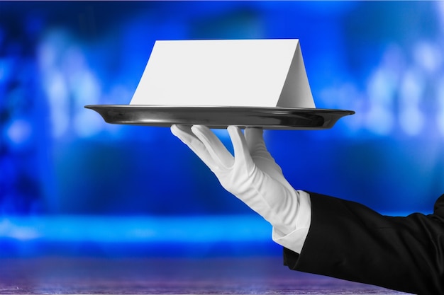 Photo waiter or butler wearing a tuxedo holding a note card on a silver tray in front of his torso.