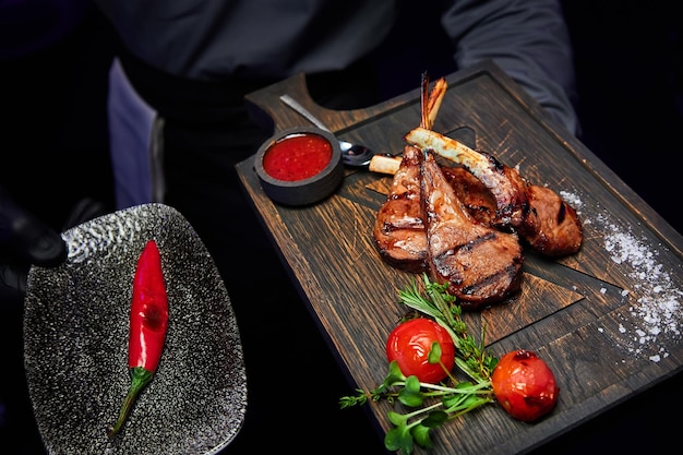 The waiter holds a dish with rack of New Zealand lamb with sauce tomatoes herbs and red hot peppers