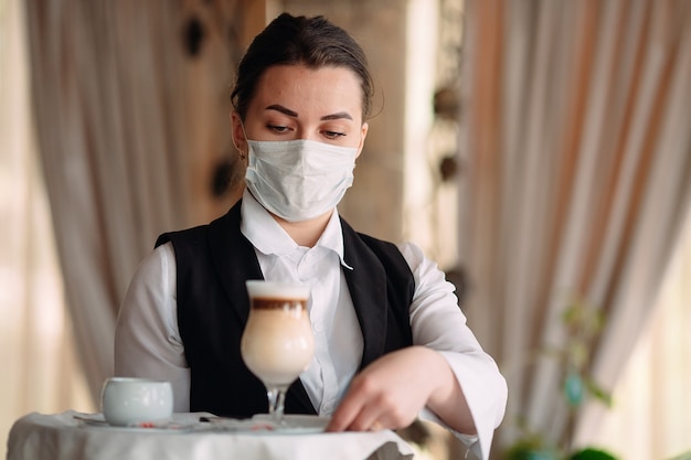 Waiter in a medical mask serves Latte coffee