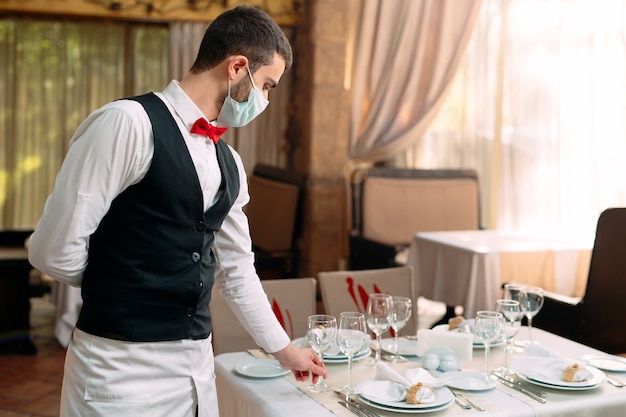 A waiter in a medical protective mask serves the table in the restaurant.