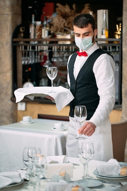 A waiter in a medical protective mask serves the table in the restaurant.