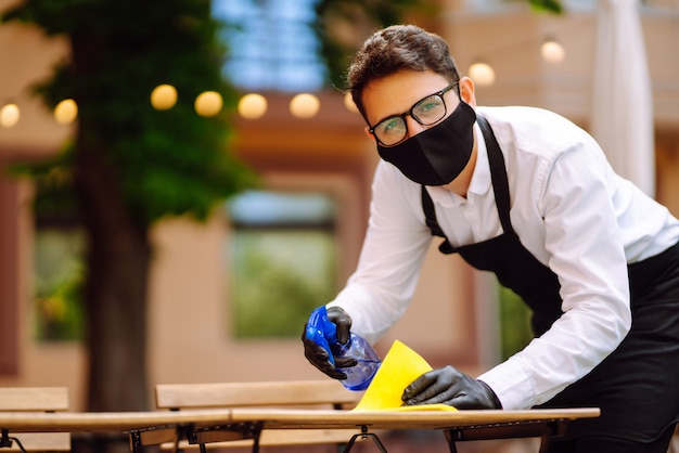 Waiter in protective mask and gloves disinfecting restaurant table for next customer.