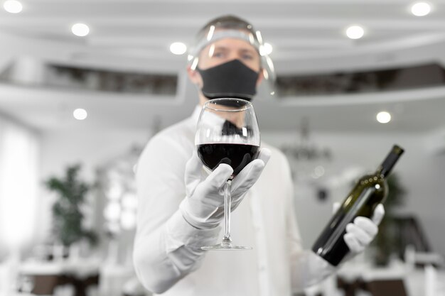 Cameriere in maschera protettiva con un bicchiere di vino rosso in mano.