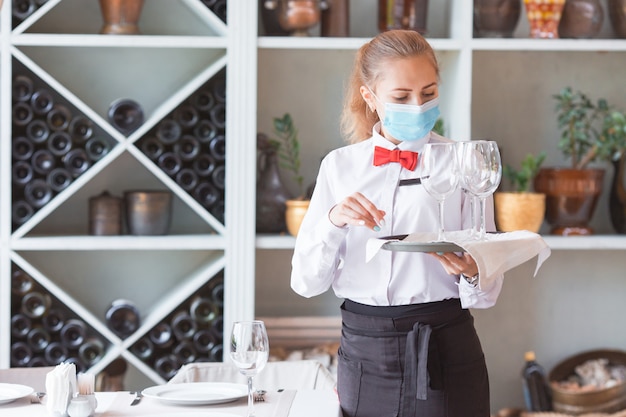 The waiter serves a table in a cafe in a protective mask.