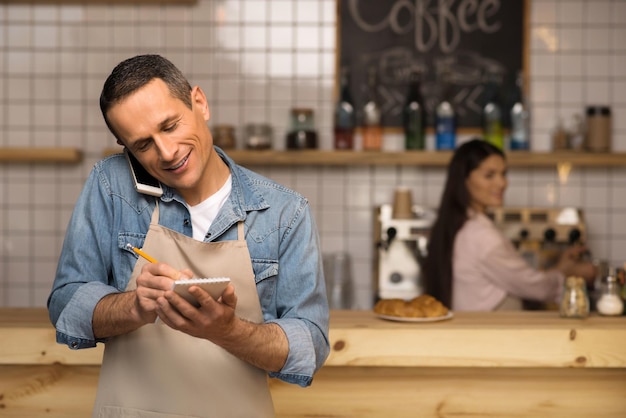 Cameriere che prende appunti e parla sullo smartphone con la cameriera sullo sfondo