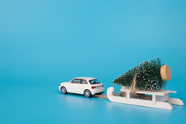크리스마스 컨셉을 기다리고 있습니다. 빈 공간이 있는 눈으로 격리된 파란색 배경에 장난감 나무가 있는 나무 흰색 미니 썰매를 운반하는 장난감 자동차의 후면 사진