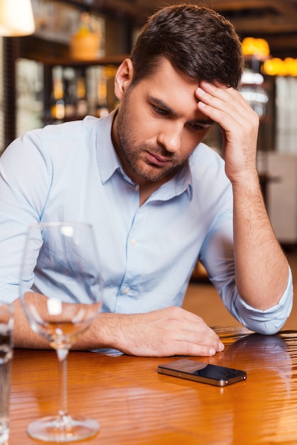 Waiting for her call. Frustrated young man holding head in hand and looking at the mobile phone while sitting at the restaurant